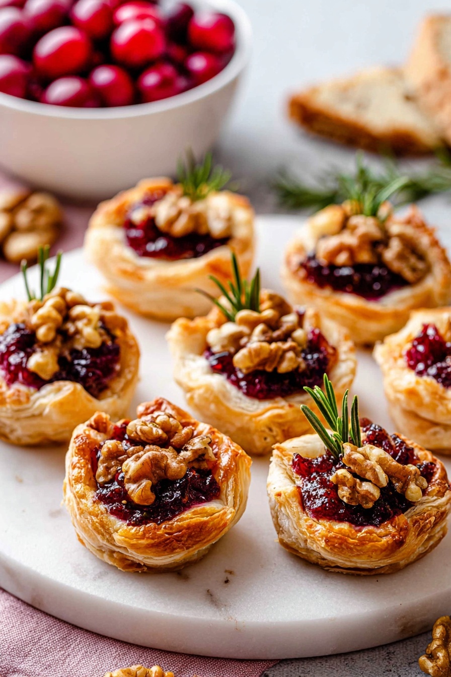 The image shows seven small puff pastry tarts arranged on a round white marbled platter. Each tart has a golden-brown, flaky pastry base with edges folded up to hold chunky dark red cranberry sauce in the center. On top of the sauce, there are small pieces of light brown walnuts and a sprig of fresh green rosemary for garnish. In the background, there is a white basket filled with bright red cranberries and a partial view of a baguette on the left side. The surface beneath the platter is a white marbled texture. photo taken with an iphone --ar 2:3 --v 7
