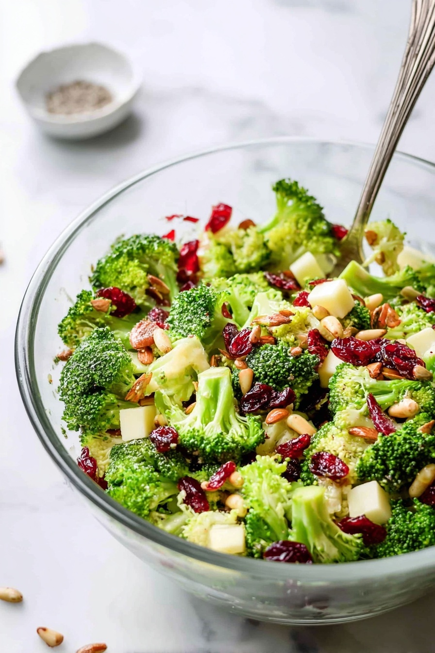 A clear glass bowl filled with a fresh broccoli salad, showing three main layers: bright green broccoli florets with a firm, bumpy texture at the bottom, topped by chunks of light yellow cheese cubes, scattered red dried cranberries, and halved almonds, all mixed evenly with sunflower seeds sprinkled throughout. The salad looks crisp and colorful, set on a white marbled surface with a silver fork resting inside the bowl. In the blurred background, a white bowl with coarse salt and cracked pepper can be seen, along with some scattered dried cranberries. Photo taken with an iphone --ar 2:3 --v 7
