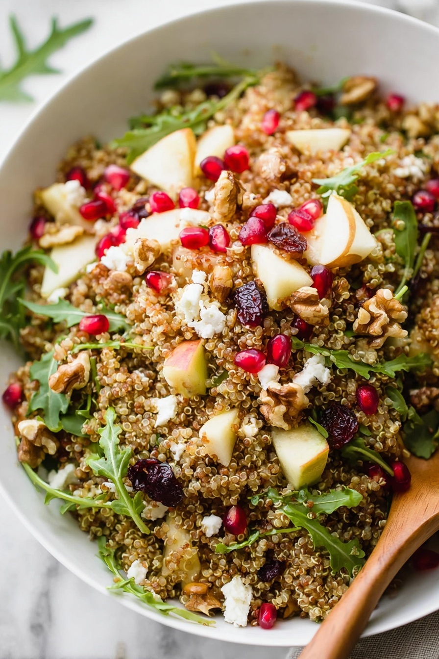A white bowl filled with a quinoa salad that has visible small beige quinoa grains as the base layer. On top, there are scattered small red pomegranate seeds, white crumbles of cheese, light yellow cubes of apple, green arugula leaves, and some darker brown nuts. A wooden spoon rests inside the bowl, slightly mixing the salad. Around the bowl, there are some pieces of quinoa, green arugula leaves, and a pomegranate cut into sections showing bright red seeds. The bowl is placed on a white marbled surface with a light, textured cloth nearby. Photo taken with an iphone --ar 2:3 --v 7