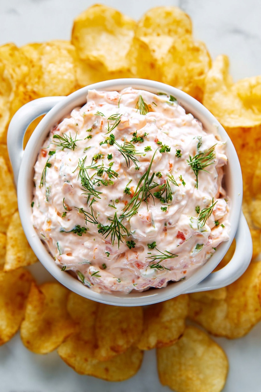 A white bowl with two handles is filled with a thick, creamy pink dip that has small green herbs mixed throughout and a few small red pieces visible inside. The surface of the dip is swirled and topped with chopped green chives and sprigs of fresh dill scattered evenly. The bowl sits on a white marbled surface surrounded by a layer of thin, round, golden-yellow potato chips that are slightly overlapping each other. Photo taken with an iphone --ar 2:3 --v 7