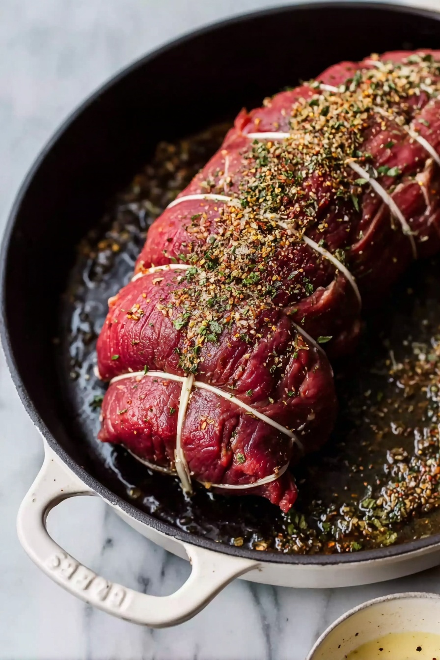 The image shows a curled raw beef roast tied with white strings in an oval shape inside a white cast iron pan. The meat is bright red with a rough texture, and it is sprinkled with dried herbs and coarse salt on top. The pan is placed on a white marbled surface, with a small white bowl containing a pale sauce seen at the bottom right corner. The scene is simple with a focus on the raw meat and seasoning, and the lighting highlights the texture and details clearly. photo taken with an iphone --ar 2:3 --v 7