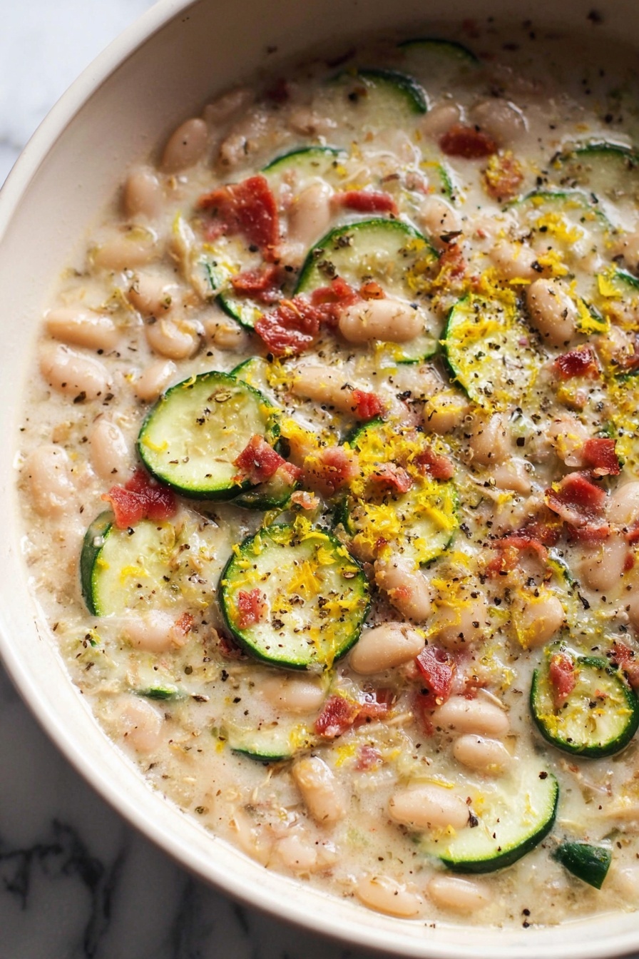 A close-up view of a creamy, thick soup in a large white pot with a dark rim. The soup has several layers of ingredients including small white beans, thin rounds of green zucchini, and small red bacon pieces scattered evenly throughout. There are visible specks of black pepper and herbs dotting the surface, with a light drizzle of oil adding shine. Thin shreds of yellow zest are sprinkled on top, giving the dish extra color and texture. The background is a white marbled surface. photo taken with an iphone --ar 2:3 --v 7