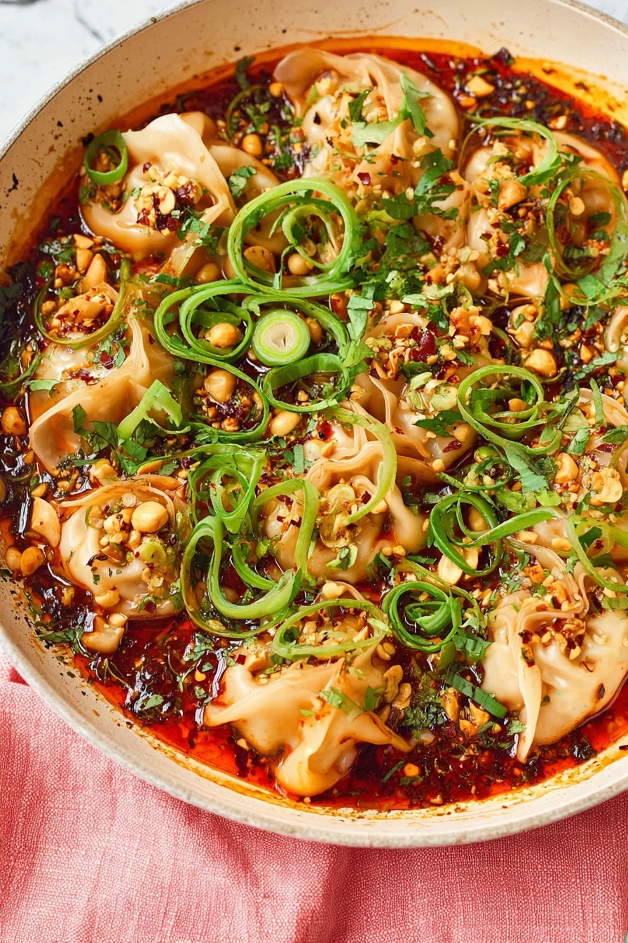 A white round pot filled with folded dumplings layered evenly on top, all covered in a thick bright red-orange sauce with oily shine and dark chili flakes. The dumplings are soft and white with some brown edges from cooking. Green herbs and sliced green onions are scattered over the top, along with crushed nuts adding crunchy texture. A woman's hand holds a gold spoon scooping some dumplings from the right side. The pot sits on a pink cloth on a white marbled surface, with a white plate with green dots and wooden chopsticks below the pot, slightly out of focus. Photo taken with an iphone --ar 2:3 --v 7