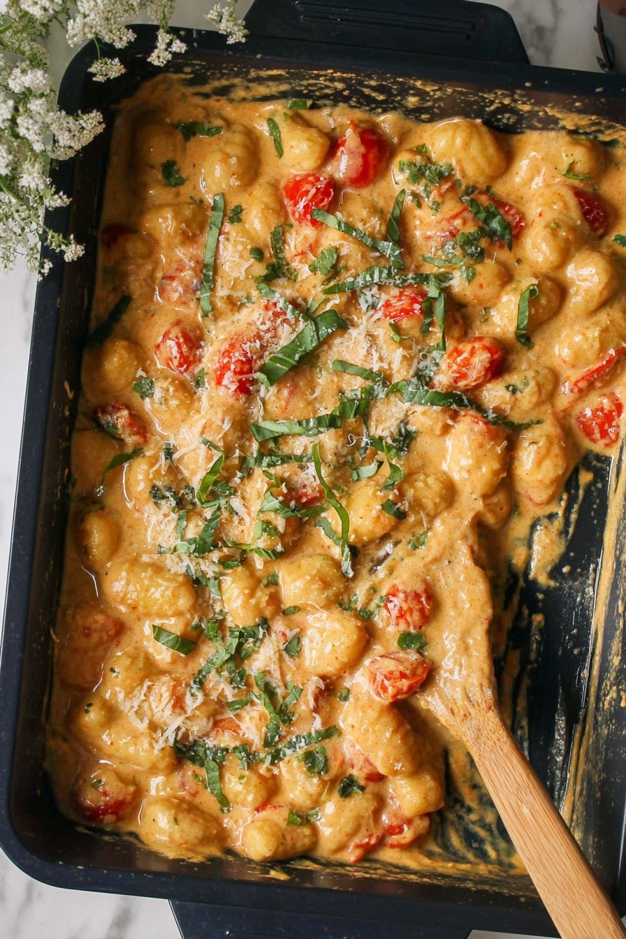 A close-up top view of a baking tray filled with creamy gnocchi mixed with small cherry tomatoes and garnished with chopped fresh green basil leaves and some grated cheese on top, showing a rich yellow-orange sauce that coats the gnocchi evenly; a wooden spatula is resting on the right side inside the tray, all placed on a white marbled surface photo taken with an iphone --ar 2:3 --v 7