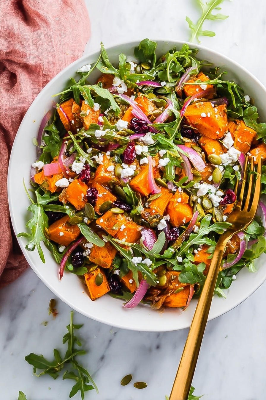 A white bowl filled with a colorful salad is shown on a white marbled surface. The salad has three main layers: bright orange chunks of roasted sweet potato spread on top, fresh green arugula leaves mixed throughout, and small white crumbles of cheese sprinkled over the vegetables. Thin strips of red onion are scattered in between, adding a hint of purple, along with small dried red cranberries and green pumpkin seeds for texture. A shiny gold fork rests inside the bowl on the right side, and a soft pink cloth is partly visible on the bottom left edge. photo taken with an iphone --ar 2:3 --v 7