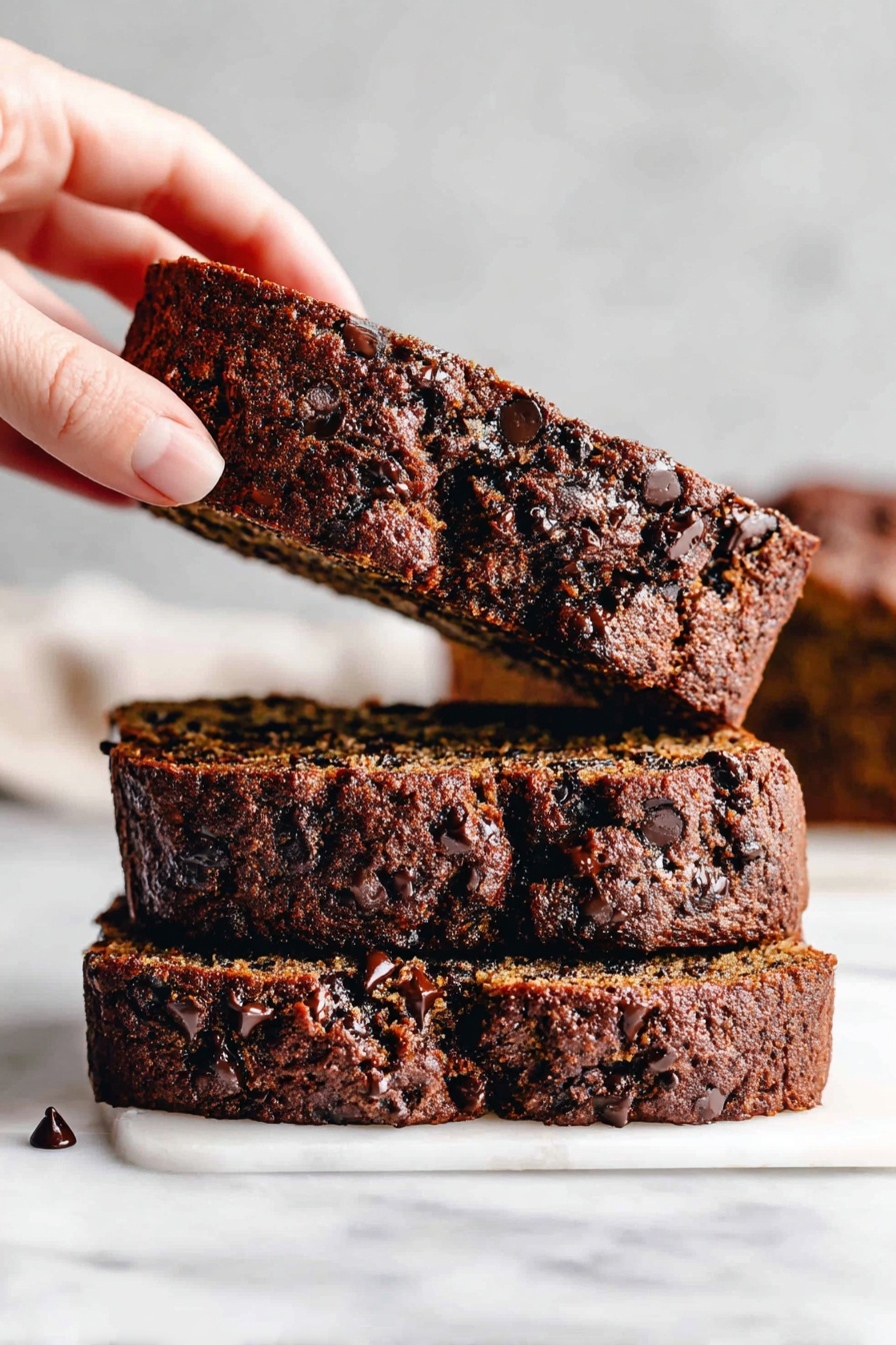 Three thick slices of dark brown chocolate chip bread stacked on a white marbled surface, with the top slice being lifted by a woman's hand from the left side of the frame. The bread has a moist texture with many small chocolate chips spread throughout, and the crust is slightly darker and rough with visible chocolate chips embedded in it. The background is blurred, showing more slices stacked together. Photo taken with an iphone --ar 2:3 --v 7