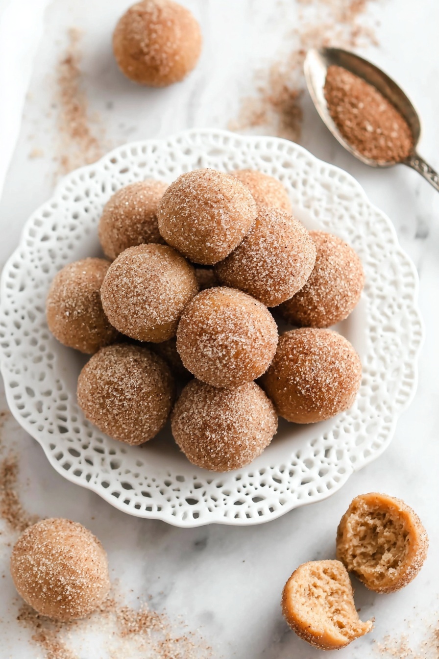 A pile of round, golden brown doughnut holes covered with a fine layer of cinnamon sugar sits stacked in the center of a white plate with a lace-like edge design. The doughnut holes have a textured, slightly rough surface from the sugar coating, and one is broken open in front of the plate, showing a soft, light beige inside with a fluffy texture. Nearby, a silver spoon holds extra cinnamon sugar, and a glass of a light brown drink is blurred in the background, all set on a white marbled surface. photo taken with an iphone --ar 2:3 --v 7