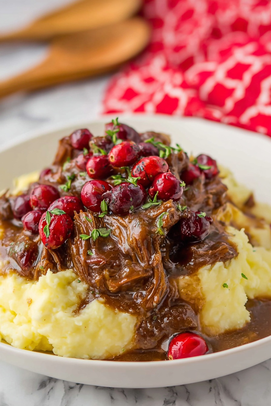 A white round plate on a white marbled surface holds a layered meal with three main parts. On the left side, there is a base layer of creamy mashed potatoes that are pale yellow and smooth. On top of the mashed potatoes, there is a thick layer of dark brown shredded meat with a rich brown sauce, scattered with red cranberries and garnished with small bits of fresh green herbs. To the right of the mashed potatoes and meat, there are about ten bright green asparagus stalks neatly aligned, showing a slightly charred texture. In the background, there is a small white bowl filled with more red cranberries. The plate is placed on a red tartan cloth with green, yellow, and blue stripes, and this rests on a wooden cutting board. A wooden spoon with a woman's hand holding it is seen blurred behind the plate. Photo taken with an iphone --ar 2:3 --v 7