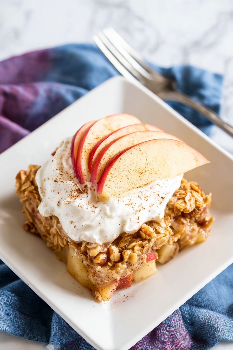 A square white plate holds a round baked oatmeal topped with a thick white cream dollop sprinkled with light brown cinnamon. Beneath the cream, thin red and yellow apple slices fan out. The oatmeal base shows chunky apple pieces, toasted oats, and walnut halves mixed within, creating a chunky texture in warm beige and orange tones. The plate rests on a white marbled surface with a folded purple and blue cloth napkin nearby and a shiny silver fork placed partially on the napkin. Photo taken with an iphone --ar 2:3 --v 7