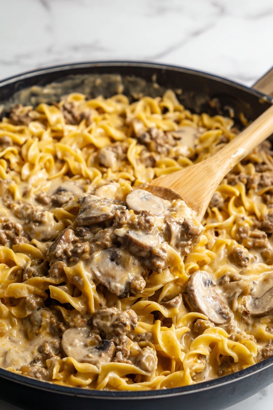 This image shows a close-up of creamy beef and mushroom pasta in a black skillet. The dish has two main layers: the bottom layer is the cooked egg noodles, light yellow and soft with a slight curl, and the top layer is a creamy sauce mixed with browned ground beef and sliced mushrooms, light beige with brown tones from the beef and the slightly darker mushrooms. A wooden spoon is lifting a scoop from the skillet, showing a mixture of noodles, beef, and mushrooms coated in the creamy sauce. The background has a white marbled texture. Photo taken with an iphone --ar 2:3 --v 7