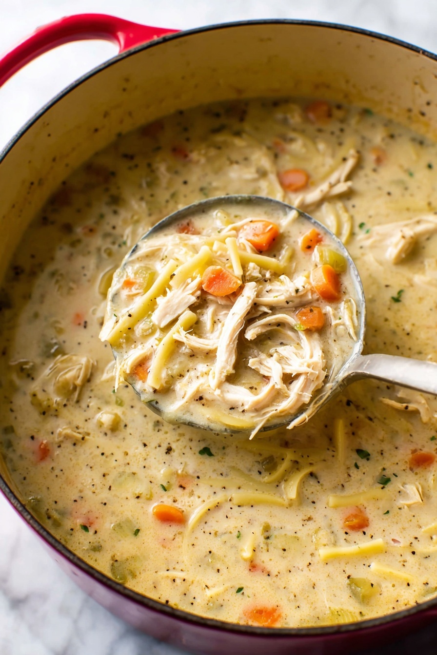 A close-up image of thick chicken noodle soup in a red pot with a beige inner surface. The soup has creamy yellow broth filled with light brown shredded chicken pieces, thick flat noodles, thin orange carrot slices, and small green celery slices mixed with black pepper specks. A metal ladle holds some soup above the pot, showing all ingredients inside the ladle clearly, with the ladle handle extending upwards. The scene is set on a white marbled texture. photo taken with an iphone --ar 2:3 --v 7