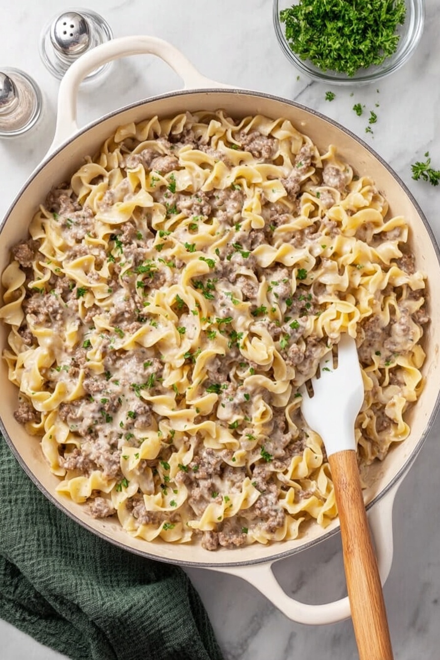 A white skillet filled with a creamy pasta dish made of egg noodles and ground meat, coated in a light beige sauce with small chopped green herbs sprinkled on top. A white spoon with a wooden handle is partially inside the skillet, slightly lifting some of the pasta. The skillet sits on a white marbled surface with a salt shaker nearby and a patterned towel in the corner. The dish looks rich and comforting with mixed textures from the tender noodles and ground meat. photo taken with an iphone --ar 2:3 --v 7