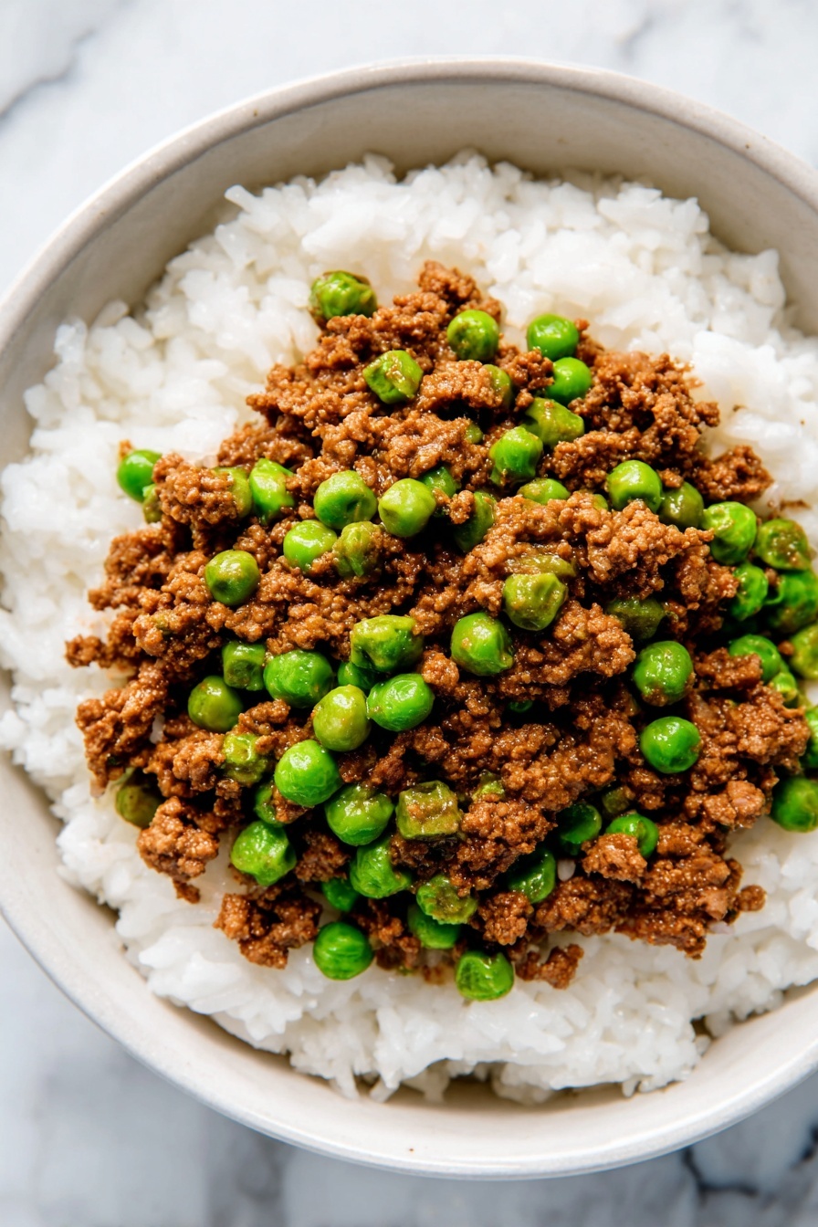 The dish shows two main layers on a white plate with a light brown rim, placed on a white marbled surface. The bottom layer is white cooked rice, soft and fluffy with visible grains clumped together. On top is a layer of cooked ground meat, brown in color with a crumbly texture, mixed evenly with bright green peas that add a pop of color. The meat and peas layer is spread thickly and covers most of the rice beneath. Photo taken with an iphone --ar 2:3 --v 7