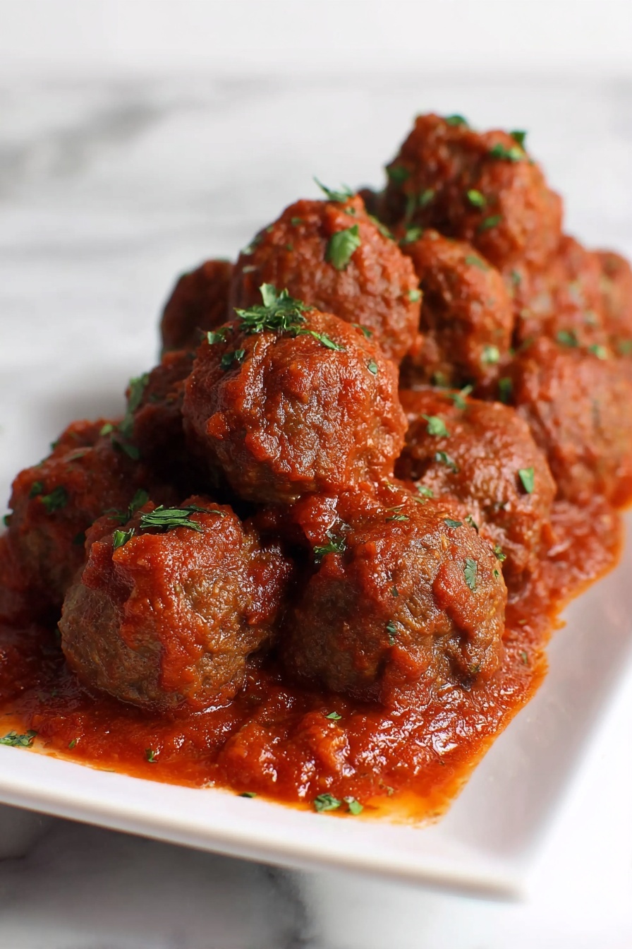 A white rectangular plate holds multiple brown meatballs covered in thick red tomato sauce, with small pieces of green herbs sprinkled on top of the meatballs and sauce. The sauce pools slightly around the base of the meatballs on the plate, creating a rich texture contrast against the smooth white plate and white marbled surface background. Photo taken with an iphone --ar 2:3 --v 7