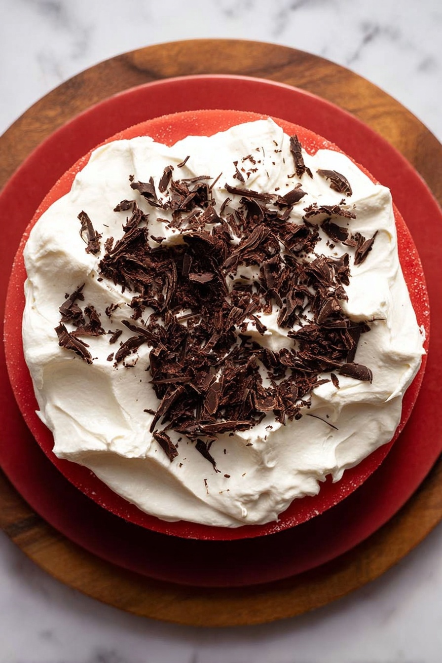 The image shows a cake with three layers. The bottom layer is dark brown and looks like a firm cookie or chocolate base. The middle layer is thick, bright red, smooth, and glossy, sitting evenly above the base. The top layer is a thick, fluffy white cream spread unevenly with a soft texture. On top of the cream, there are thin dark brown chocolate shavings scattered around. The cake is placed on a wooden round board with a natural texture, and the background is a white marbled surface. Photo taken with an iphone --ar 2:3 --v 7