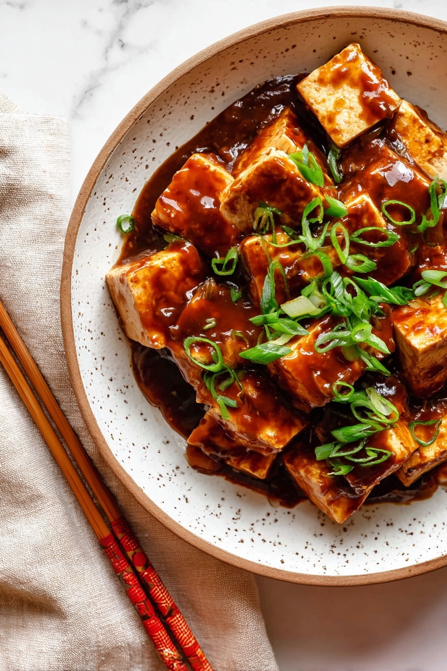 This image shows many chunks of tofu coated in a thick, shiny, dark brown sauce placed in a white bowl with a brown rim. On top, there are scattered slices of bright green spring onions, adding a fresh touch to the rich sauce. The bowl sits on a white marbled surface next to a folded light cloth and a pair of wooden chopsticks wrapped with a red and gold decoration. The overall look is warm and appetizing, with a focus on the glistening sauce and vibrant green onions. photo taken with an iphone --ar 2:3 --v 7